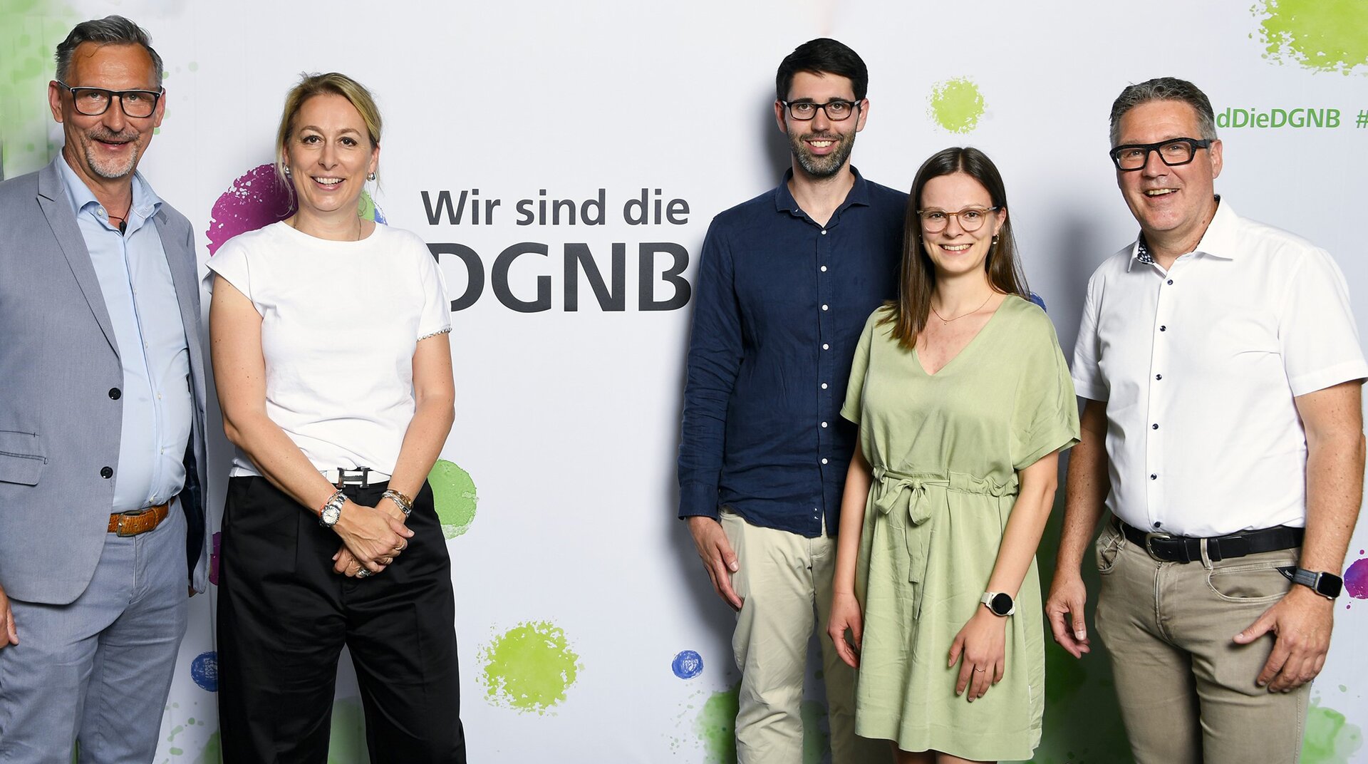 Fünf Personen stehen vor einem Banner mit der Aufschrift "Wir sind die DGNB". Zwei Männer und eine Frau stehen links, eine Frau und ein Mann stehen rechts. Alle blicken lächelnd in die Kamera.