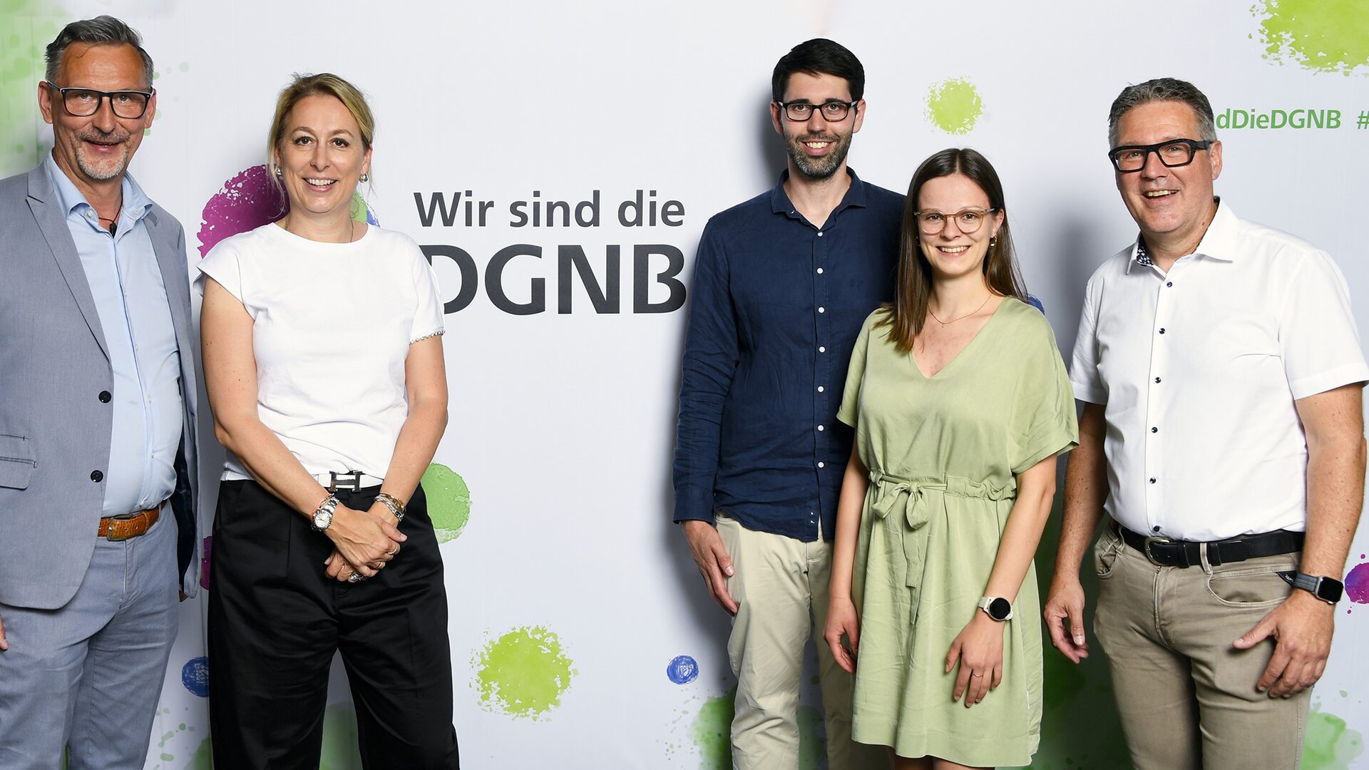 Fünf Personen stehen vor einem Banner mit der Aufschrift "Wir sind die DGNB". Zwei Männer und eine Frau stehen links, eine Frau und ein Mann stehen rechts. Alle blicken lächelnd in die Kamera.