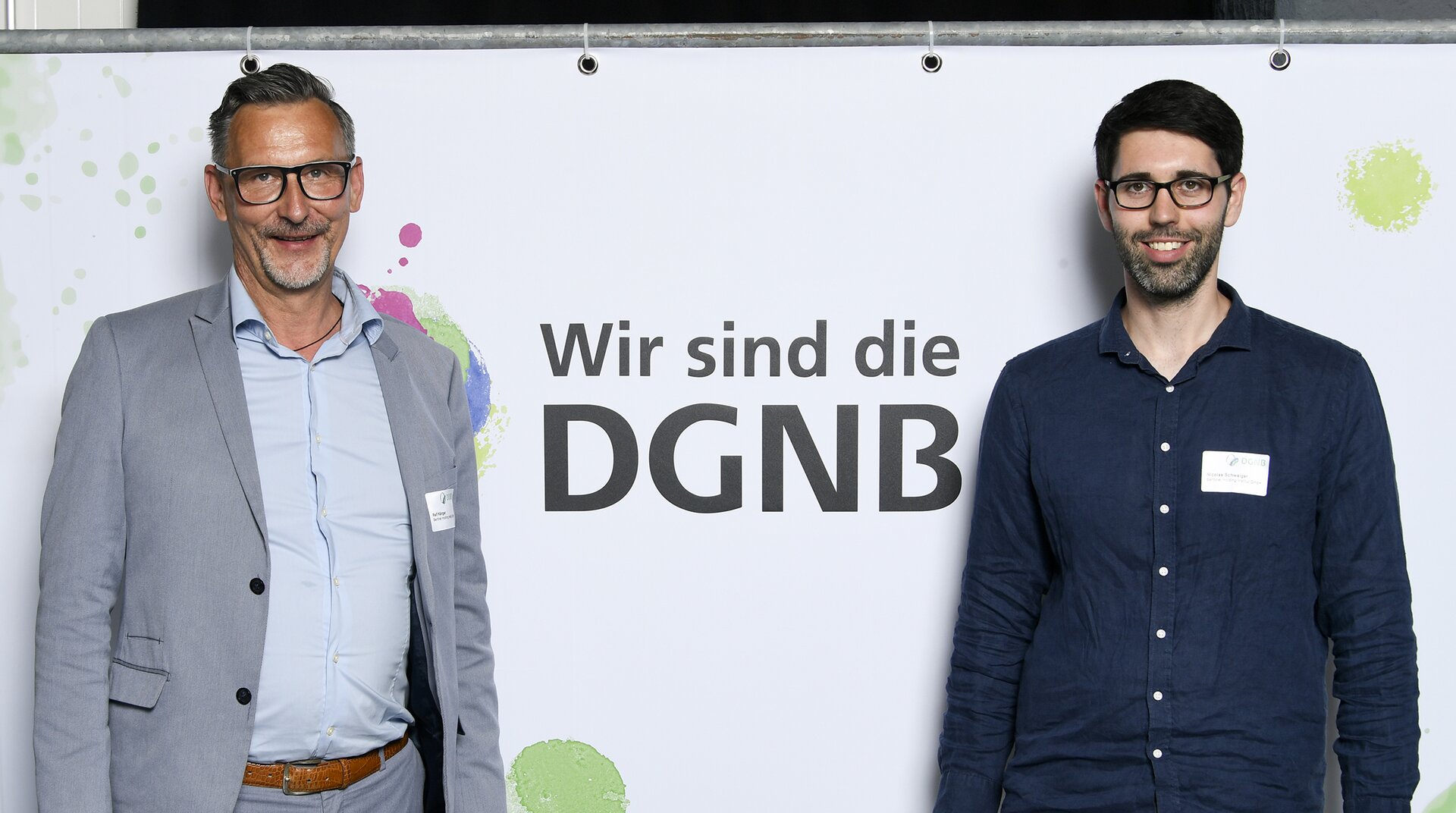 Zwei Männer stehen vor einem Banner mit der Aufschrift "Wir sind die DGNB". Der Mann links trägt einen grauen Anzug, der Mann rechts ein dunkles Hemd. Beide lächeln in die Kamera.